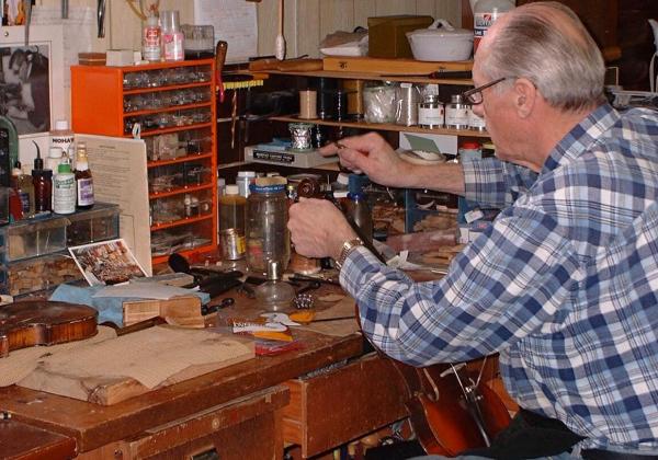 Old man in plaid shirt at a workbench tuning up a violin