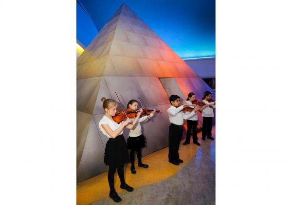 Group of children playing violin in a museum in front of a smaller scale pyramid