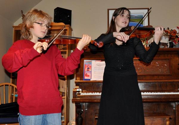 Young teen boy in a red sweater playing a violin duet with Rhiannon, clad in formal black
