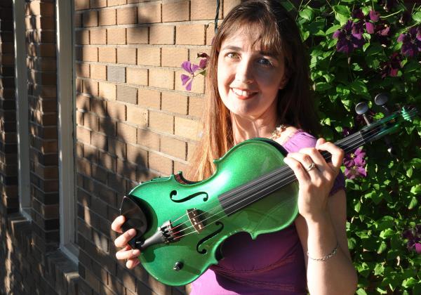 Rhiannon in purple standing in front of a brick wall with vines and purple flowers holding a green violin