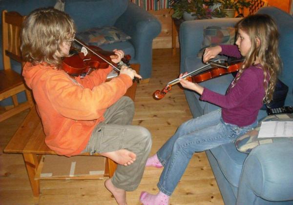 Brother and sister facing each other causally and playing violins in the livingroom