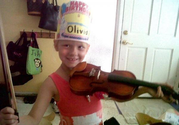 Little girl wearing a happy birthday paper crown with "Olivia" written on it and holding her new violin