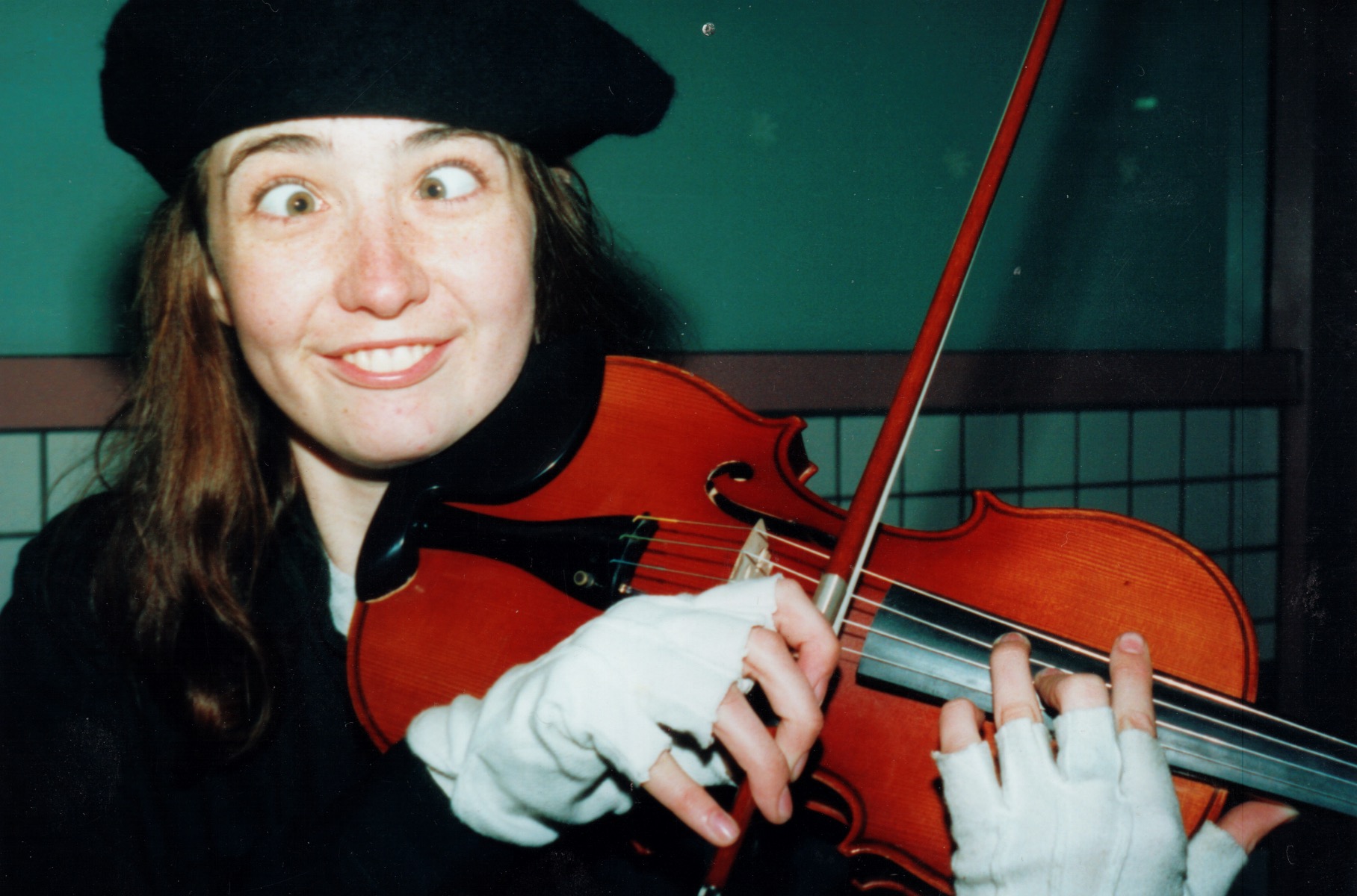 Rhiannon in a coat, playing violin with fingerless gloves, making a funny face and crossing her eyes