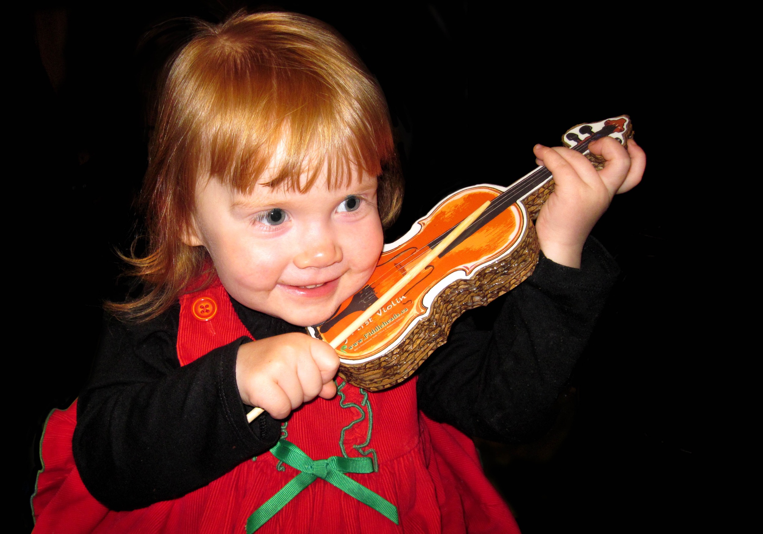 Red-headed toddler in a red dress smiling and playing a cardboard violin with a chopstick as a bow