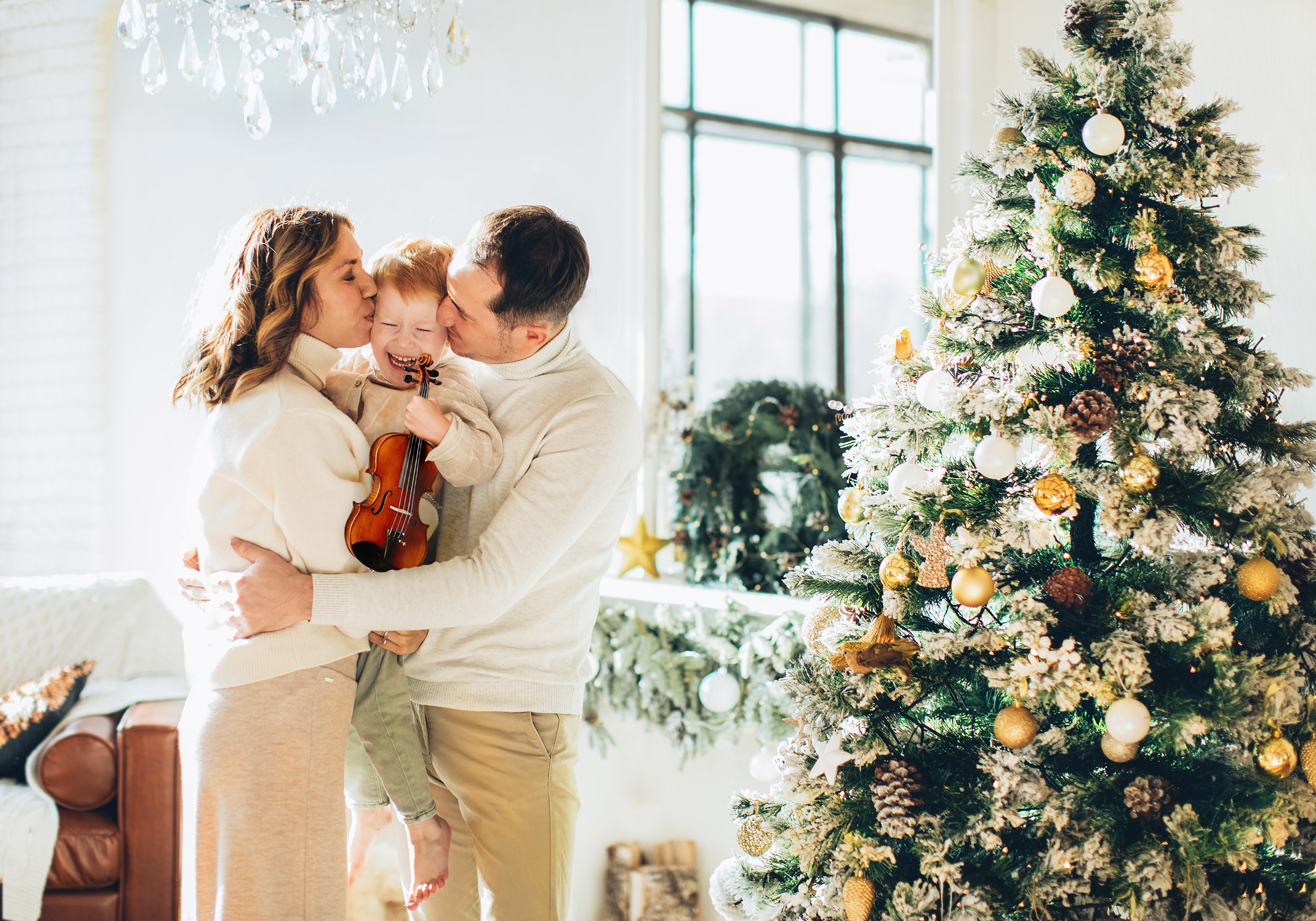 A mom and dad standing by a Christmas tree are holding and kissing their preschooler child while he smiles and holds his new violin
