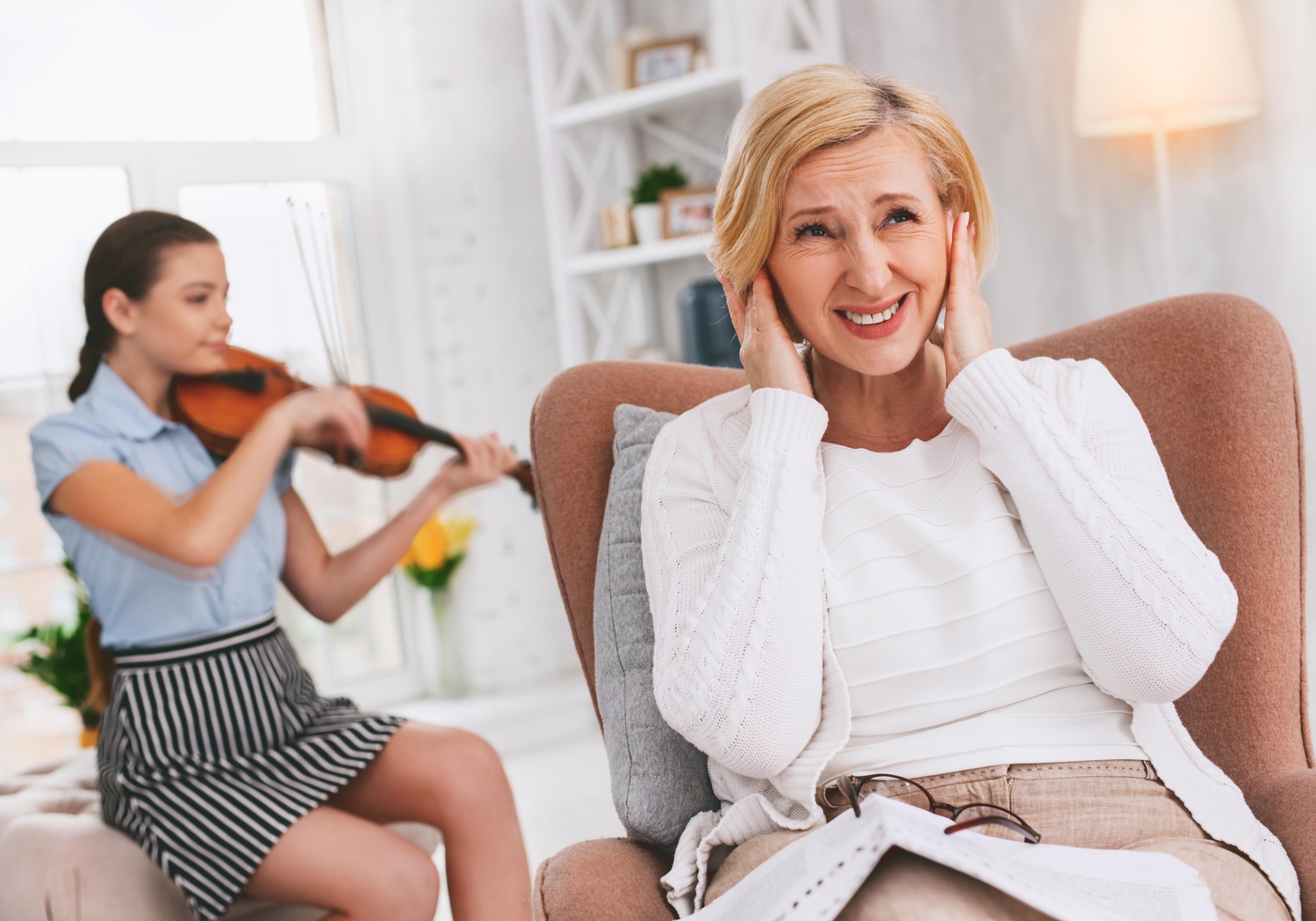 Woman with a grimace/smile and her hands over her ears while a young girl behind her plays violin