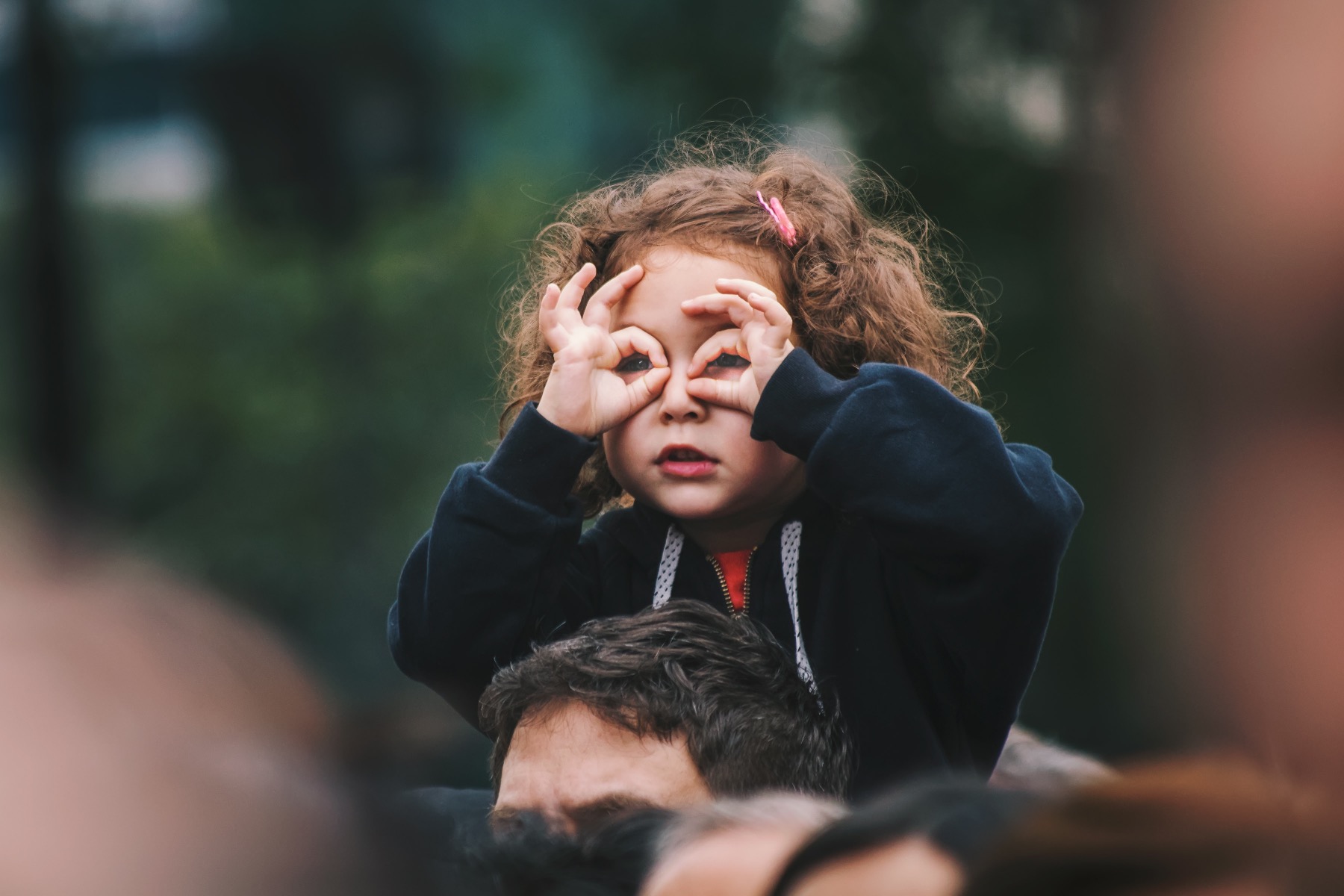 Preschool girl sitting on her dad's shoulders and making glasses with her fingers while watching a concert