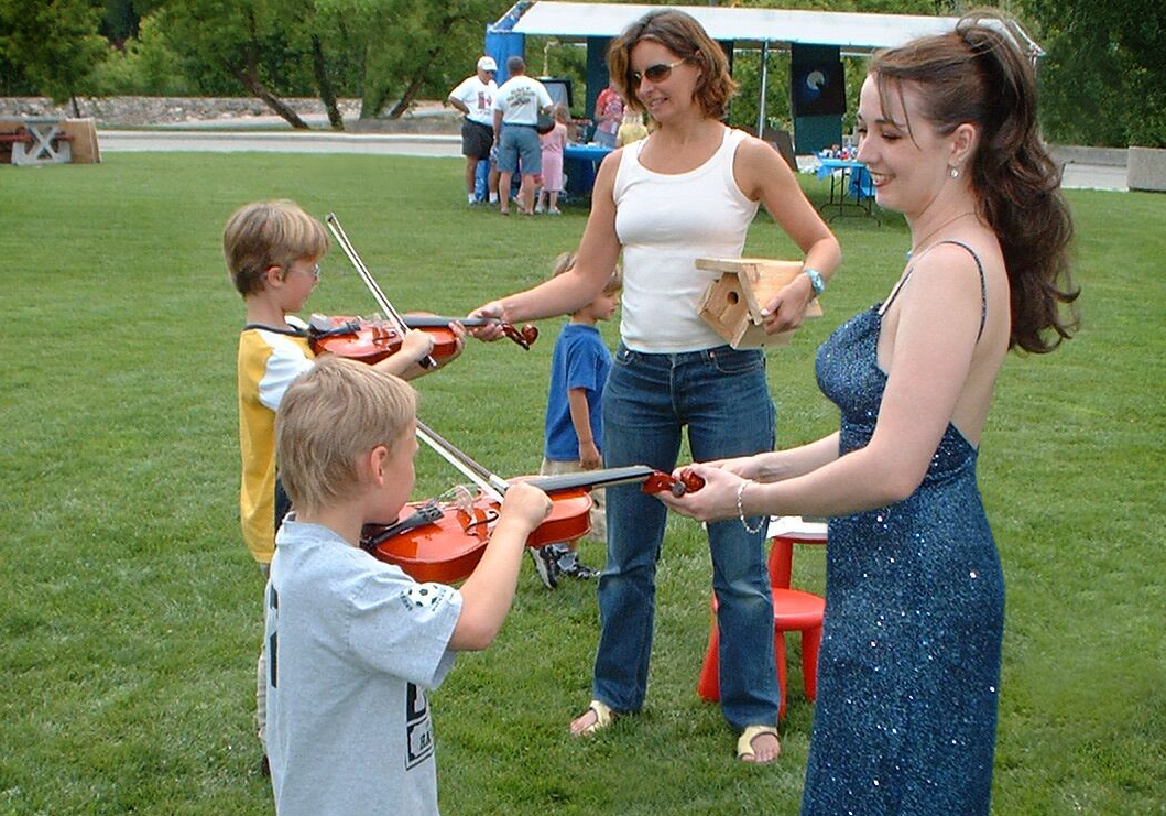Rhiannon and a mom help children try the violin for the first time in a park