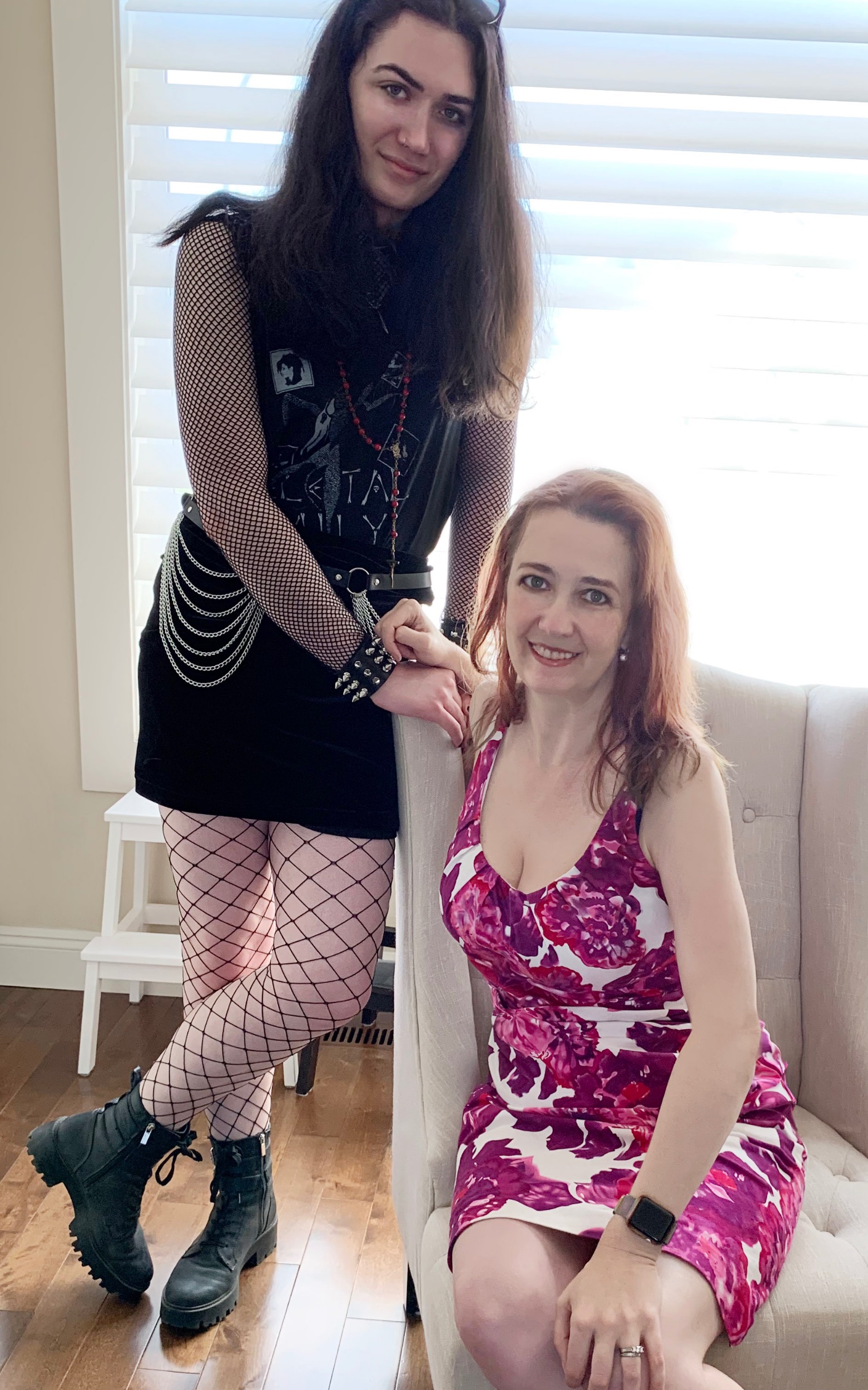 Lydia in goth clothes standing beside a seated Rhiannon in a hot pink floral dress