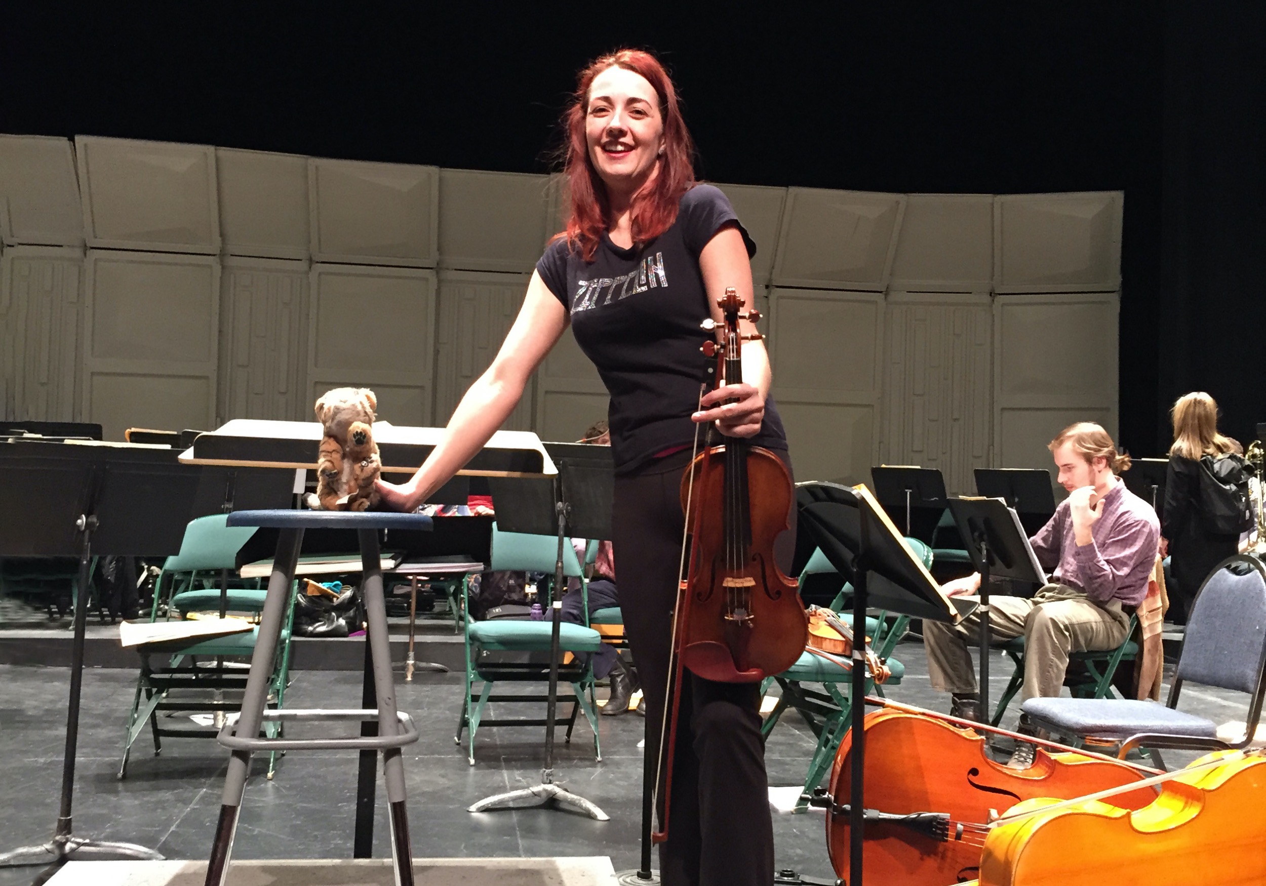 Rhiannon on stage at a symphony rehearsal in a Led Zeppelin shirt and holding a stuffed tiger on the conductor's stand