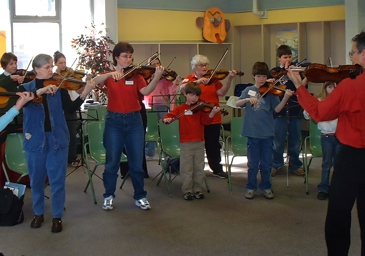 Group of violinists of all ages playing with a teacher in a red dress shirt