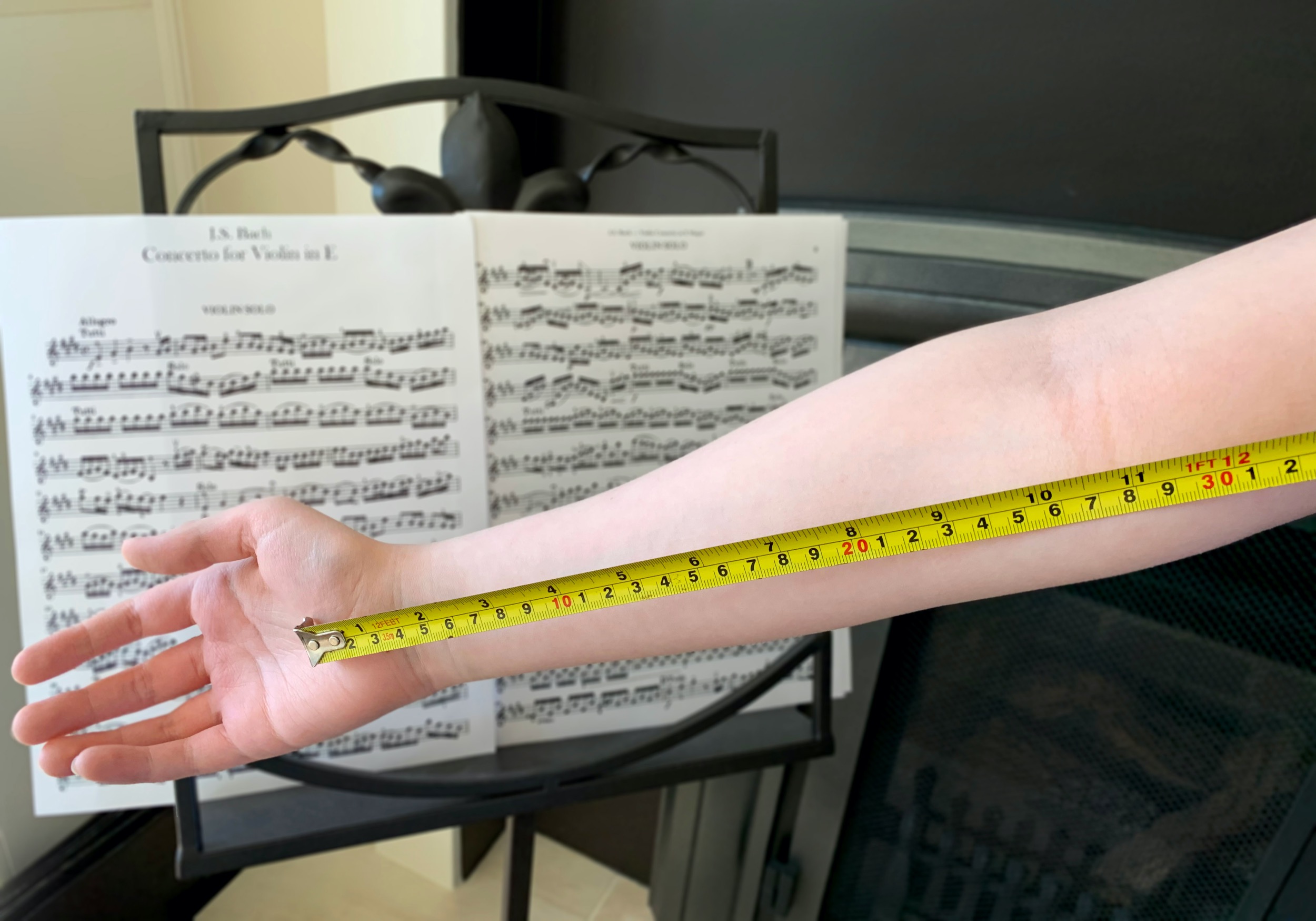 Child's arm extended with a yellow measuring tape and a music stand with Bach sheet music in the background
