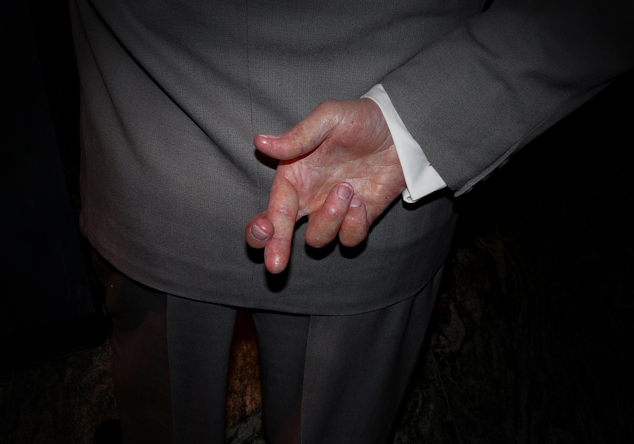 suited old man in shadows crosses his fingers behind his back
