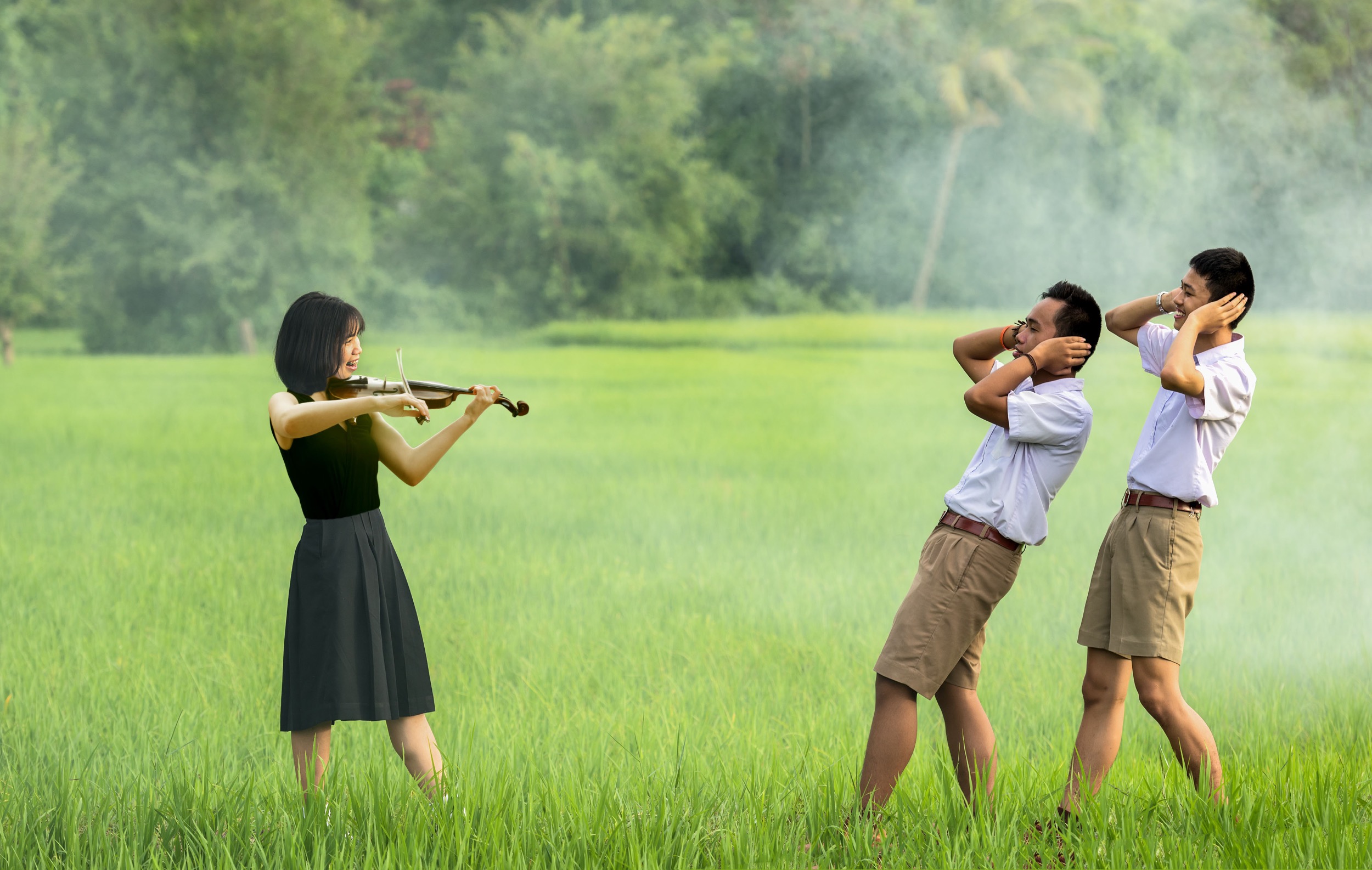 Young attractive Asian woman in a black dress playing violin in a misty green field while two fit asian men in khaki shorts cover their ears in pain