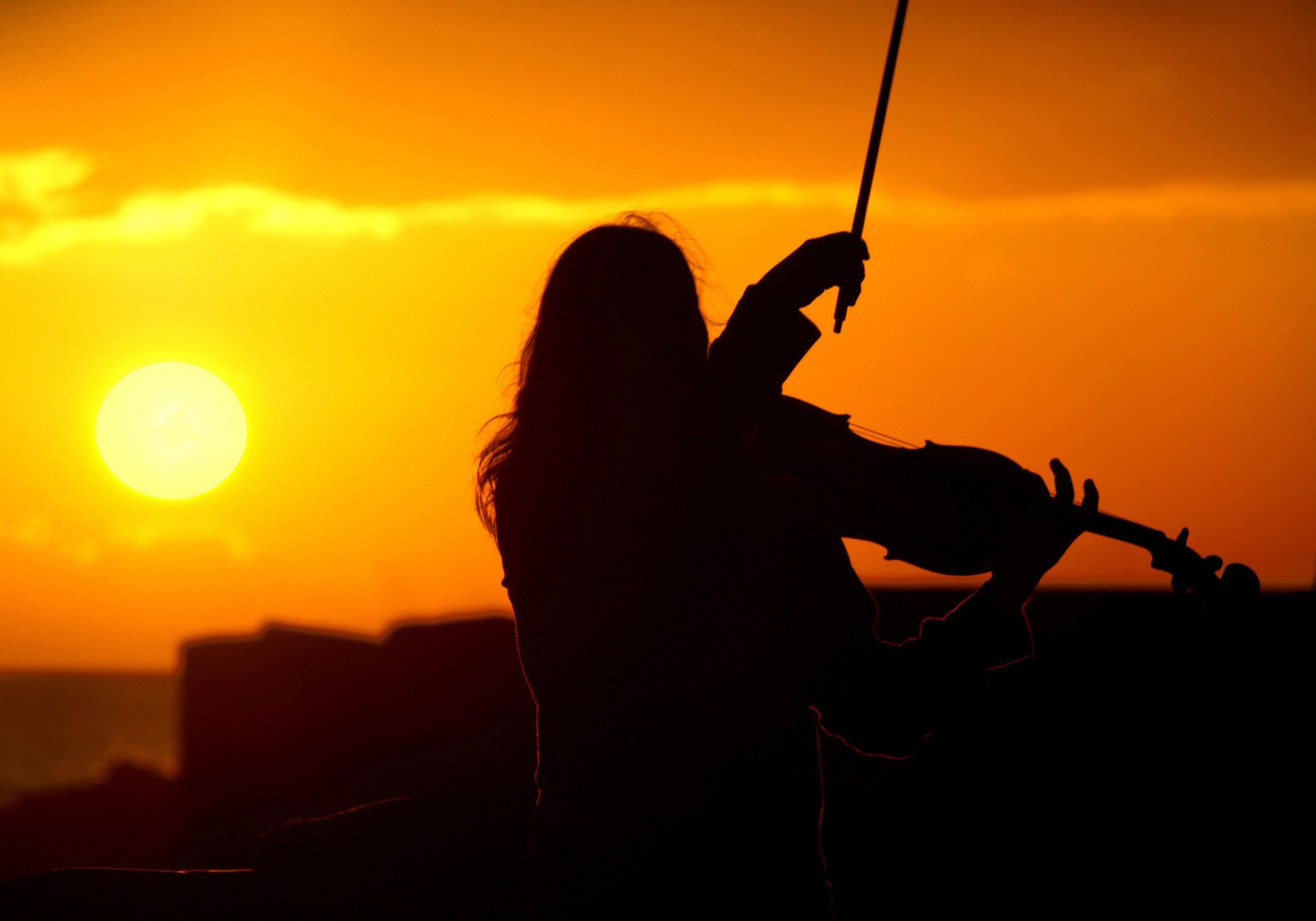 Silhouette of a woman playing violin with a fiery orange sunset and ocean behind her