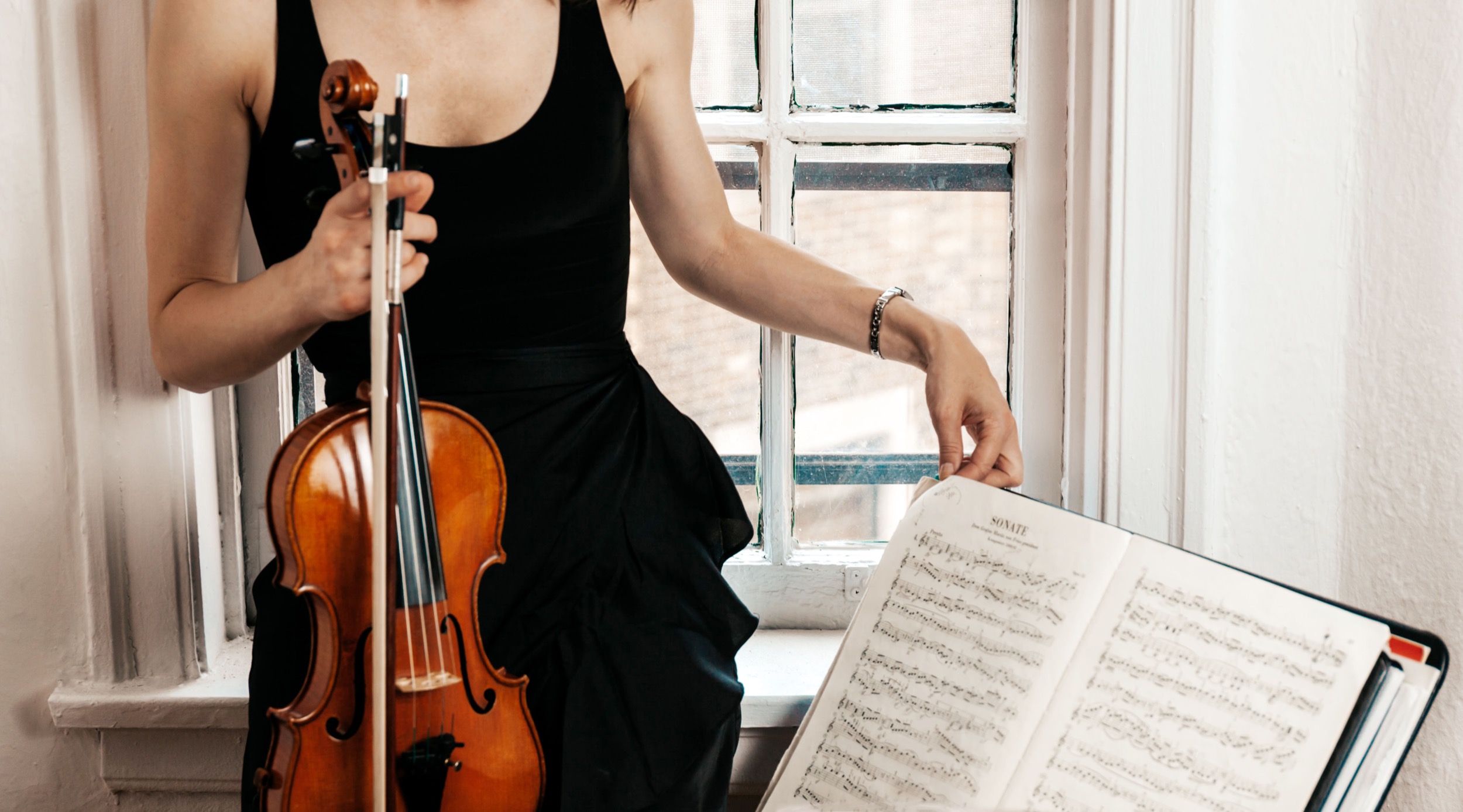 thin white woman in black dress holding her violin in front of an old painted white windowsill and preparing sheet music on a stand