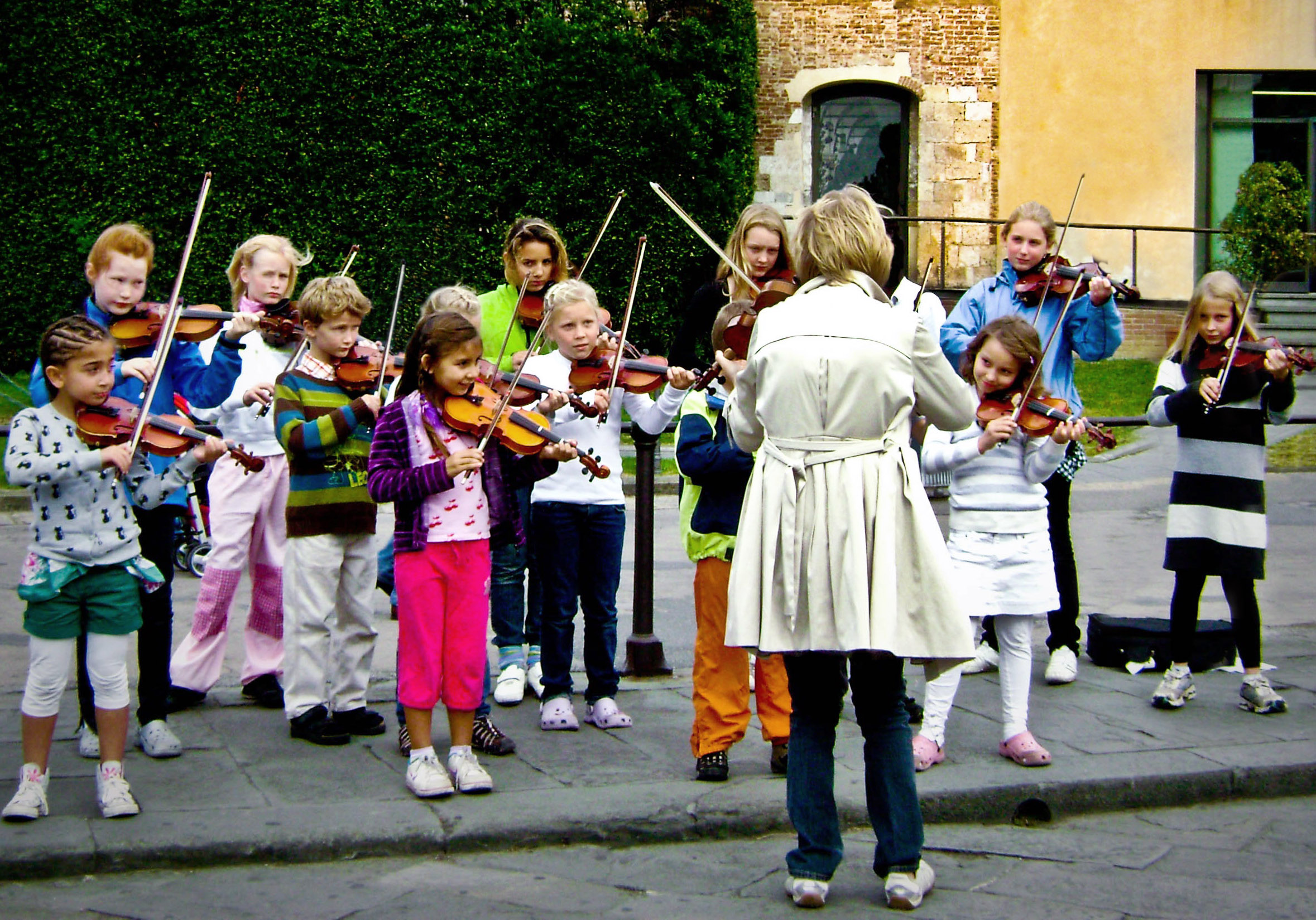 Teacher and a group of children of many ages all playing violin in front of an old brick building