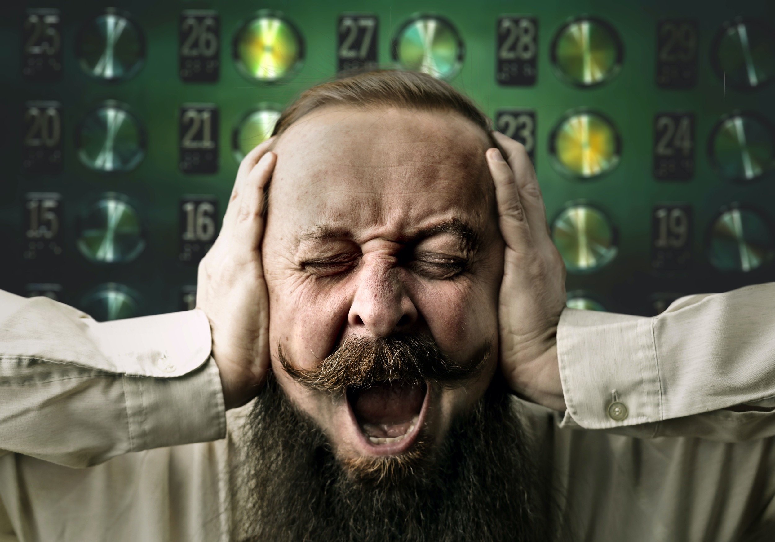 Bearded man in front of a panel of elevator buttons covering his ears and shouting in pain